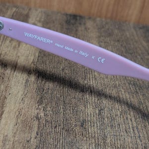 Puede incluir: Primer plano de la patilla de unas gafas de sol Wayfarer de color rosa claro. El texto "WAYFARER&reg; Hand Made in Italy K CE" est&aacute; impreso en blanco. El fondo es una superficie de madera.