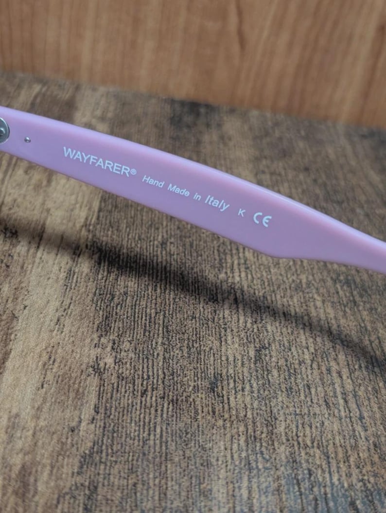 Puede incluir: Primer plano de la patilla de unas gafas de sol Wayfarer de color rosa claro. El texto "WAYFARER&reg; Hand Made in Italy K CE" est&aacute; impreso en blanco. El fondo es una superficie de madera.