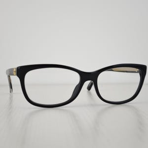 Puede incluir: Gafas negras con acabado brillante y forma de montura ojo de gato. Las patillas presentan un pequeño detalle metálico circular de tono dorado cerca de las bisagras. Los cristales transparentes están montados en una estructura de plástico.