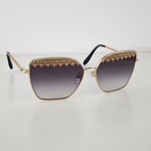 May include: Gold-tone metal sunglasses featuring a geometric frame shape and gradient gray lenses. The upper rim of each lens is accented with a decorative gold-tone lattice pattern. The thin temples have a textured finish and black tips.