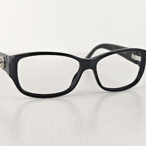 Puede incluir: Gafas rectangulares negras con acabado brillante. Las monturas presentan un emblema circular de tono plateado en la bisagra de la patilla. Los cristales transparentes están montados en una estructura de aro completo con patillas curvas.