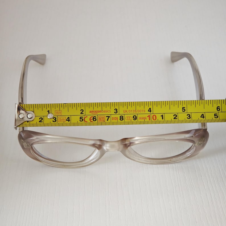 May include: Translucent taupe-colored eyeglasses with a rounded frame shape. A yellow measuring tape is positioned across the front, showing a width of approximately 5.5 inches from the outer edge of one lens frame to the other.