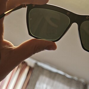 Peut inclure: Lunettes de soleil noires avec des verres vert fonc&eacute;. Les montures sont rectangulaires avec des bords arrondis et de petits accents m&eacute;talliques. Les branches sont fines et dor&eacute;es. Les lunettes sont tenues par une main.