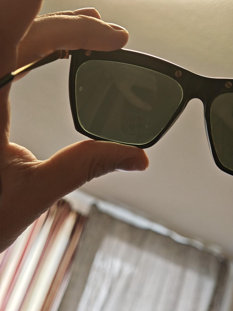 Peut inclure: Lunettes de soleil noires avec des verres vert fonc&eacute;. Les montures sont rectangulaires avec des bords arrondis et de petits accents m&eacute;talliques. Les branches sont fines et dor&eacute;es. Les lunettes sont tenues par une main.