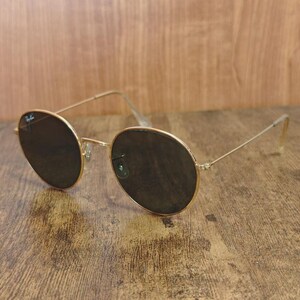 May include: Gold-framed sunglasses with dark lenses. The round lenses are set in a thin, gold metal frame. The temples are also gold and have a slight curve at the ear. The sunglasses are resting on a wooden surface.