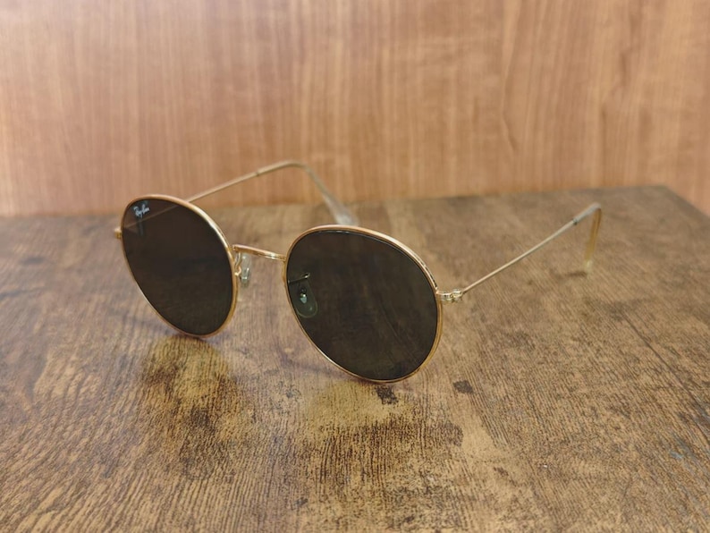 May include: Gold-framed sunglasses with dark lenses. The round lenses are set in a thin, gold metal frame. The temples are also gold and have a slight curve at the ear. The sunglasses are resting on a wooden surface.