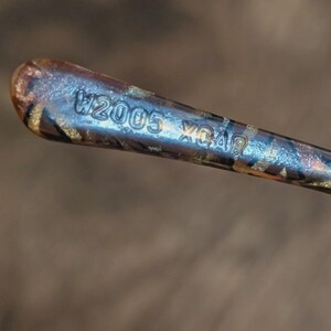 Peut inclure: Gros plan d'un objet m&eacute;tallique avec le texte "W2005 XO40" grav&eacute; sur la surface. L'objet pr&eacute;sente un motif marron et dor&eacute; avec un &eacute;clat m&eacute;tallique. L'arri&egrave;re-plan est un marron flou.