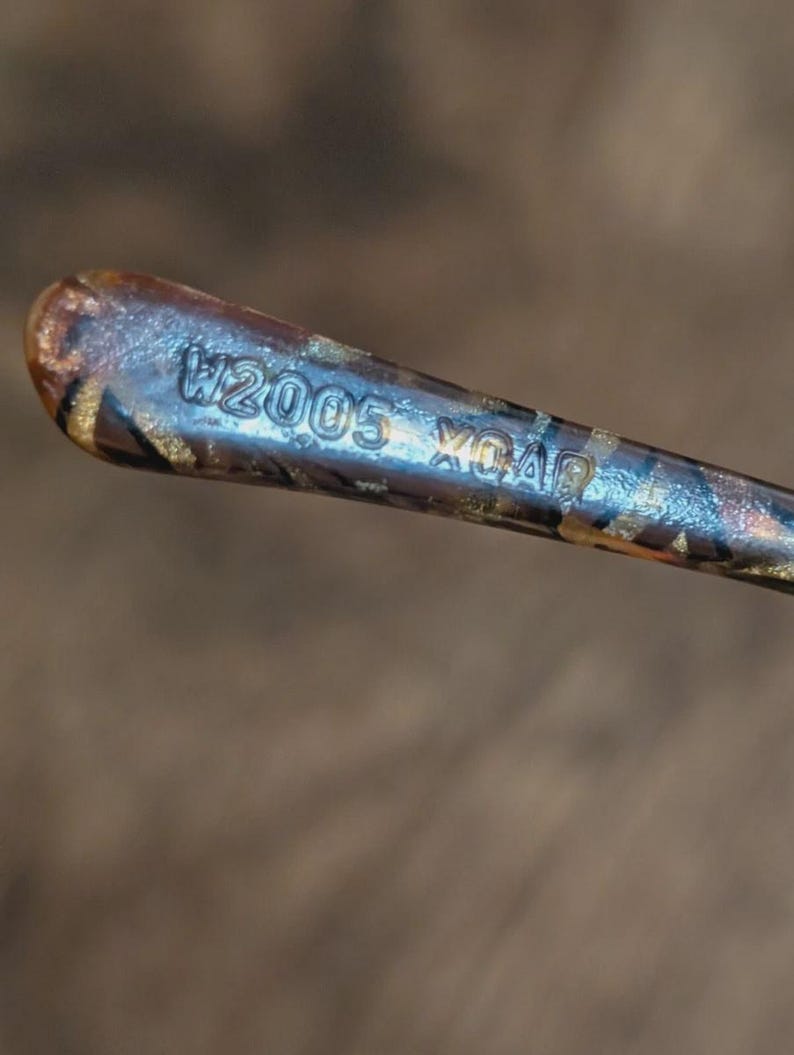 Peut inclure: Gros plan d'un objet m&eacute;tallique avec le texte "W2005 XO40" grav&eacute; sur la surface. L'objet pr&eacute;sente un motif marron et dor&eacute; avec un &eacute;clat m&eacute;tallique. L'arri&egrave;re-plan est un marron flou.