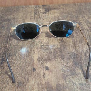 May include: Aviator-style sunglasses with a gold-colored frame and dark lenses. The sunglasses have a double bridge and curved temples. The lenses are oval-shaped and reflect the surrounding environment. The sunglasses are designed for sun protection.