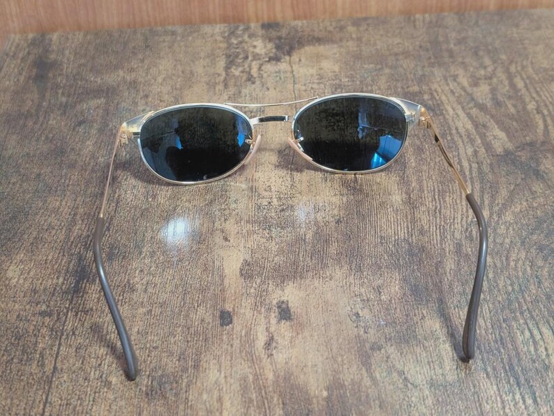 May include: Aviator-style sunglasses with a gold-colored frame and dark lenses. The sunglasses have a double bridge and curved temples. The lenses are oval-shaped and reflect the surrounding environment. The sunglasses are designed for sun protection.