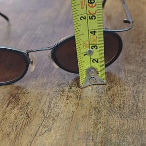 May include: Black oval sunglasses with brown lenses. The glasses are being measured with a yellow tape measure. The tape measure shows the glasses are approximately 2 inches in height.