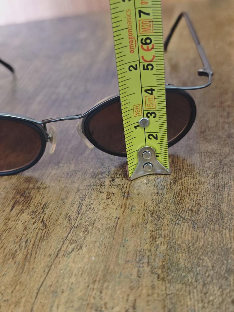 May include: Black oval sunglasses with brown lenses. The glasses are being measured with a yellow tape measure. The tape measure shows the glasses are approximately 2 inches in height.