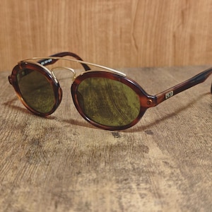 May include: Vintage sunglasses with a brown tortoiseshell frame and gold-tone metal accents. The oval lenses are a green tint. The arms are a dark brown color. The sunglasses are resting on a wooden surface.