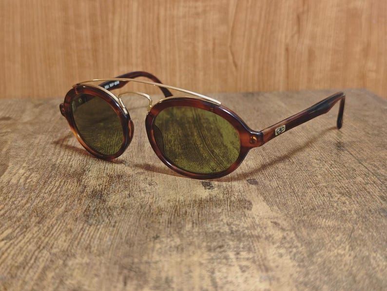 May include: Vintage sunglasses with a brown tortoiseshell frame and gold-tone metal accents. The oval lenses are a green tint. The arms are a dark brown color. The sunglasses are resting on a wooden surface.