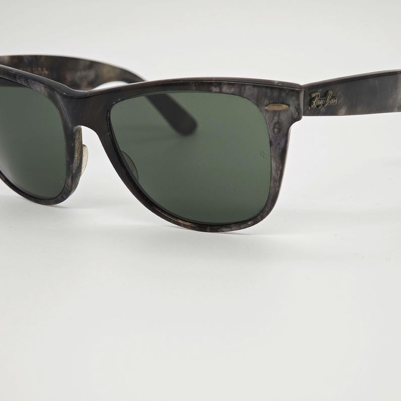 Puede incluir: Gafas de sol gris oscuro con lentes verdes. Las monturas tienen un patr&oacute;n veteado. El logotipo de Ray-Ban es visible en la patilla. Un accesorio de gafas cl&aacute;sico.