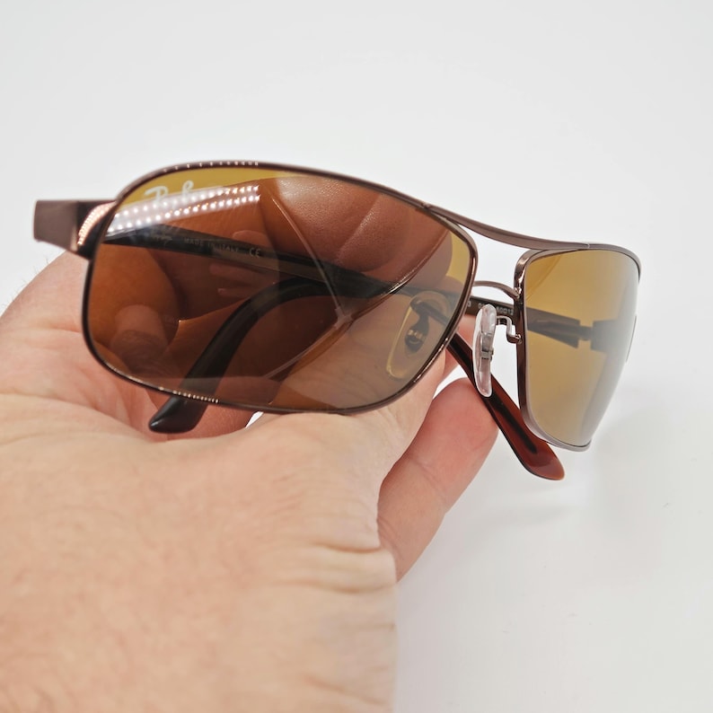May include: Brown metal sunglasses with brown tinted lenses. The sunglasses have a double bridge design and the Ray-Ban logo is visible on the top left lens. The sunglasses are held in a hand against a white background.