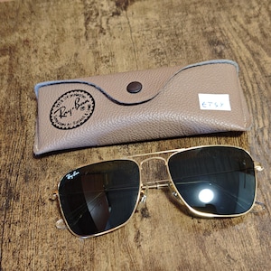 Puede incluir: Gafas de sol Ray-Ban con montura dorada y lentes oscuras y estuche a juego. El estuche es de color beige con cierre de bot&oacute;n y el logotipo de Ray-Ban. Las gafas de sol son de forma cuadrada y est&aacute;n sobre una superficie de madera.