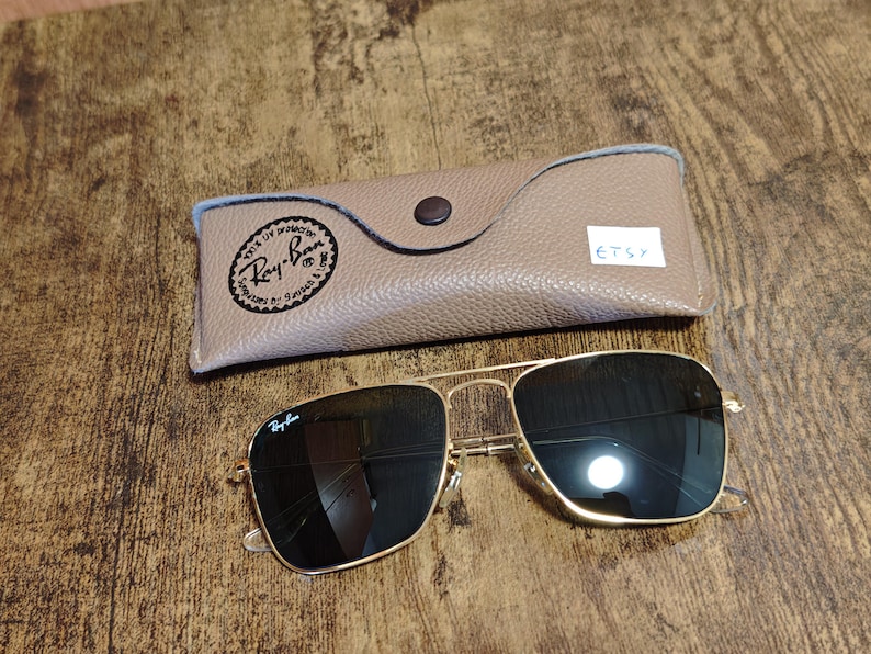 Puede incluir: Gafas de sol Ray-Ban con montura dorada y lentes oscuras y estuche a juego. El estuche es de color beige con cierre de bot&oacute;n y el logotipo de Ray-Ban. Las gafas de sol son de forma cuadrada y est&aacute;n sobre una superficie de madera.