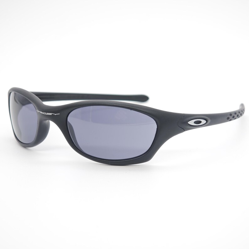 May include: Black sunglasses with gray lenses. The sunglasses have a curved frame and the Oakley logo is visible on the arm and lens. The arms have a textured design near the ear.