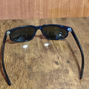May include: Dark navy sunglasses with dark lenses. The sunglasses have a classic rectangular frame and sleek, curved arms. The hinges are silver. The sunglasses are resting on a wooden surface.