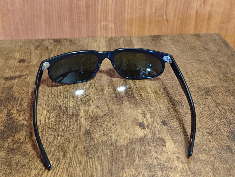 May include: Dark navy sunglasses with dark lenses. The sunglasses have a classic rectangular frame and sleek, curved arms. The hinges are silver. The sunglasses are resting on a wooden surface.