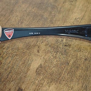 K&ouml;nnte beinhalten: Schwarzer Brillenb&uuml;gel mit dem Ducati Corse-Logo in einem rot-wei&szlig;en Schild. Der B&uuml;gel tr&auml;gt den Text "12-780 64 15" und den Markennamen "HMX".