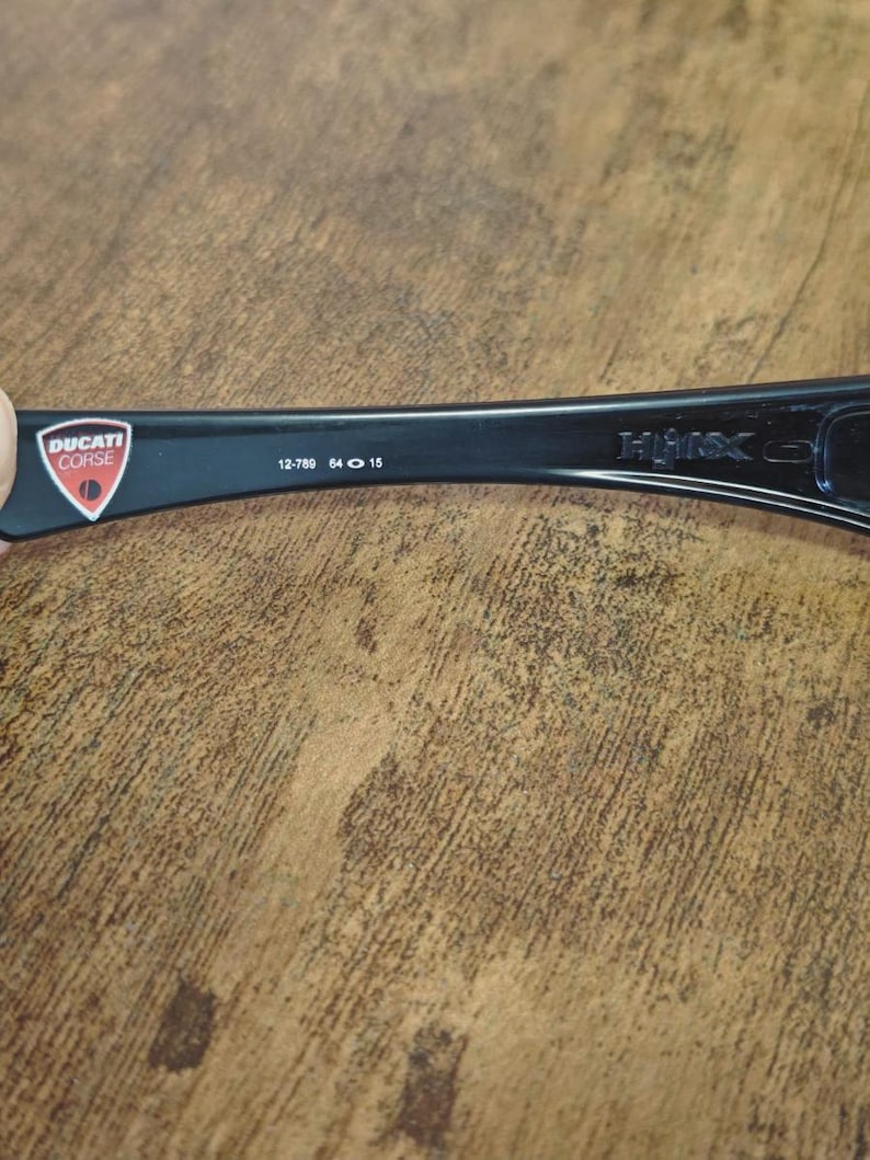 K&ouml;nnte beinhalten: Schwarzer Brillenb&uuml;gel mit dem Ducati Corse-Logo in einem rot-wei&szlig;en Schild. Der B&uuml;gel tr&auml;gt den Text "12-780 64 15" und den Markennamen "HMX".