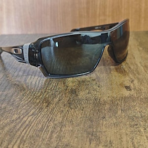 May include: Black sunglasses with a wraparound design. The sunglasses have dark lenses and a black frame with silver accents near the temples. The lenses are slightly reflective, and the sunglasses are resting on a wooden surface.