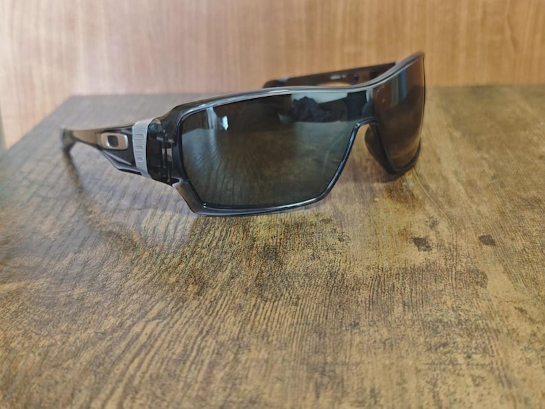 May include: Black sunglasses with a wraparound design. The sunglasses have dark lenses and a black frame with silver accents near the temples. The lenses are slightly reflective, and the sunglasses are resting on a wooden surface.