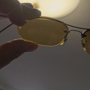 May include: Rimless sunglasses with gold-toned frames and yellow-tinted lenses. The sunglasses are held by a hand, with the Ray-Ban logo visible on the lens. The background is a soft, out-of-focus light.