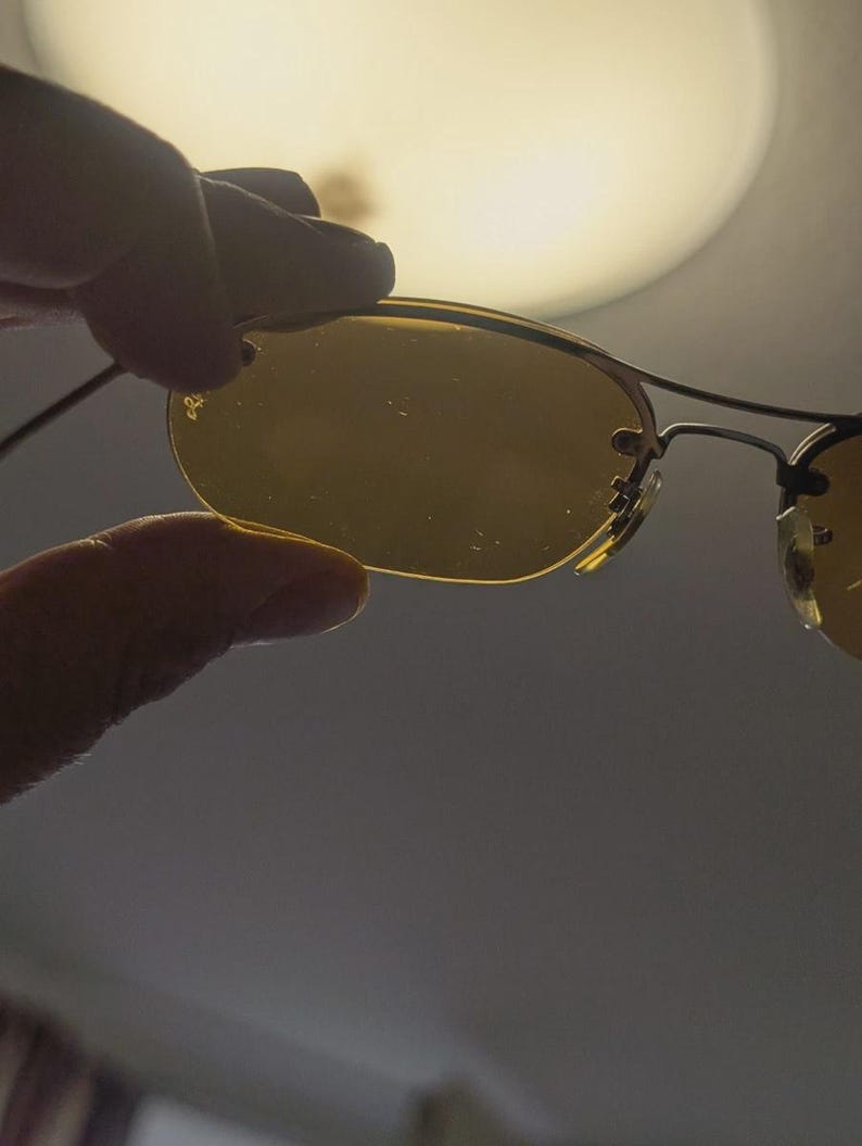 May include: Rimless sunglasses with gold-toned frames and yellow-tinted lenses. The sunglasses are held by a hand, with the Ray-Ban logo visible on the lens. The background is a soft, out-of-focus light.