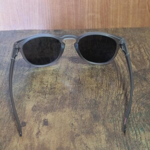 May include: Gray-framed sunglasses with dark lenses. The round frames and temples are a translucent gray color. The lenses are dark, providing shade. The sunglasses are resting on a wooden surface.
