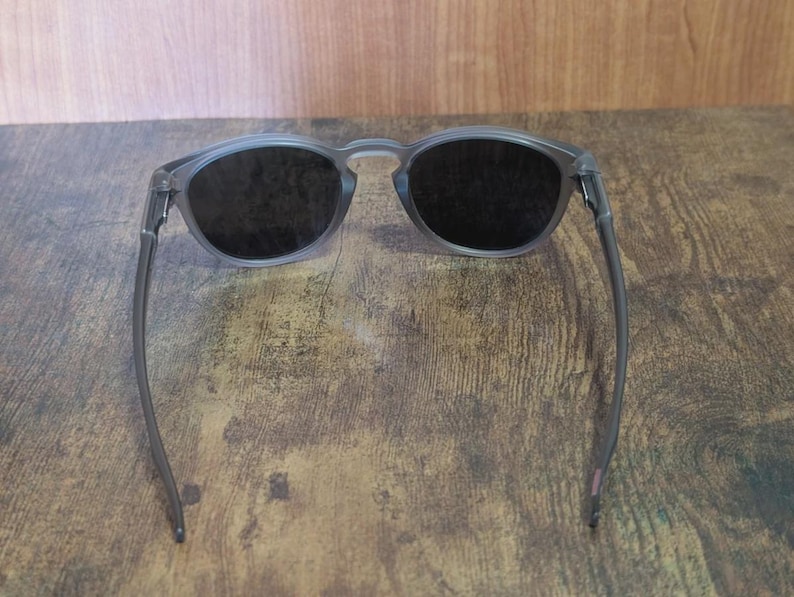 May include: Gray-framed sunglasses with dark lenses. The round frames and temples are a translucent gray color. The lenses are dark, providing shade. The sunglasses are resting on a wooden surface.