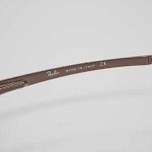 May include: Close-up of the arm of a pair of brown Ray-Ban sunglasses. The text "Ray-Ban" is visible, along with "MADE IN ITALY" and "CE". The frame is a solid, matte brown color.