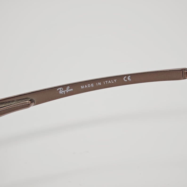 May include: Close-up of the arm of a pair of brown Ray-Ban sunglasses. The text "Ray-Ban" is visible, along with "MADE IN ITALY" and "CE". The frame is a solid, matte brown color.
