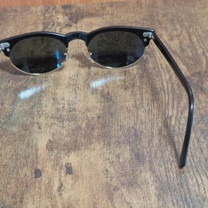 May include: Black sunglasses with a classic design. The frames are black with a gold-colored trim around the lenses. The lenses are dark, and the arms are thin and sleek. The sunglasses are resting on a wooden surface.