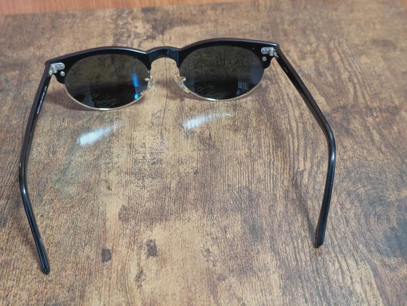 May include: Black sunglasses with a classic design. The frames are black with a gold-colored trim around the lenses. The lenses are dark, and the arms are thin and sleek. The sunglasses are resting on a wooden surface.