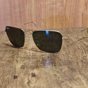 Puede incluir: Gafas de sol con montura dorada y lentes oscuras. Las gafas de sol tienen un dise&ntilde;o cl&aacute;sico de aviador con doble puente y un logotipo sutil en la lente superior izquierda. Las patillas son finas y se extienden hasta las patillas.