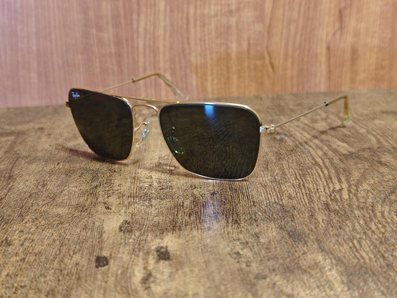 Puede incluir: Gafas de sol con montura dorada y lentes oscuras. Las gafas de sol tienen un dise&ntilde;o cl&aacute;sico de aviador con doble puente y un logotipo sutil en la lente superior izquierda. Las patillas son finas y se extienden hasta las patillas.