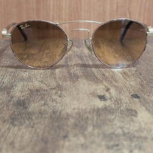 Peut inclure: Lunettes de soleil rondes avec une monture en m&eacute;tal dor&eacute; et des verres d&eacute;grad&eacute;s marron. Les lunettes ont un double pont et le logo Ray-Ban est visible sur le verre. Elles reposent sur une surface en bois.