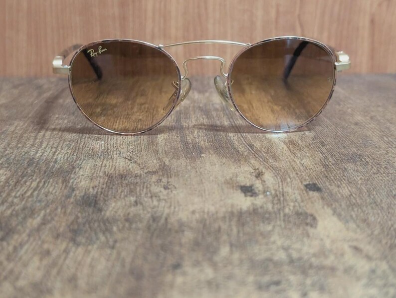 Peut inclure: Lunettes de soleil rondes avec une monture en m&eacute;tal dor&eacute; et des verres d&eacute;grad&eacute;s marron. Les lunettes ont un double pont et le logo Ray-Ban est visible sur le verre. Elles reposent sur une surface en bois.