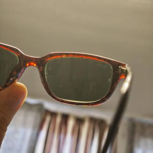 May include: A pair of tortoiseshell sunglasses with dark lenses. The frames have a reddish-brown pattern. The sunglasses are held up, with the lenses facing the viewer. The background is blurred.