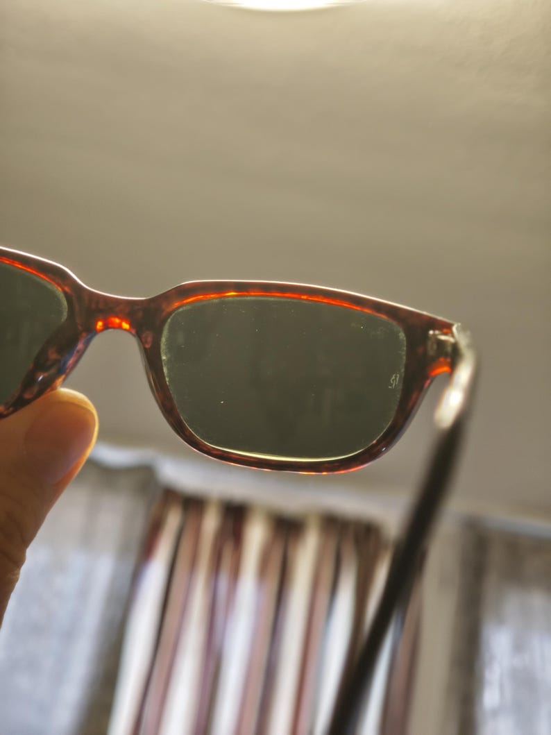 May include: A pair of tortoiseshell sunglasses with dark lenses. The frames have a reddish-brown pattern. The sunglasses are held up, with the lenses facing the viewer. The background is blurred.
