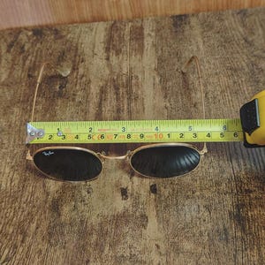 May include: Gold-framed sunglasses with dark lenses, measuring approximately 5.5 inches in width. The Ray-Ban logo is visible on the left lens. The glasses are set against a wooden background, with a measuring tape indicating the size.