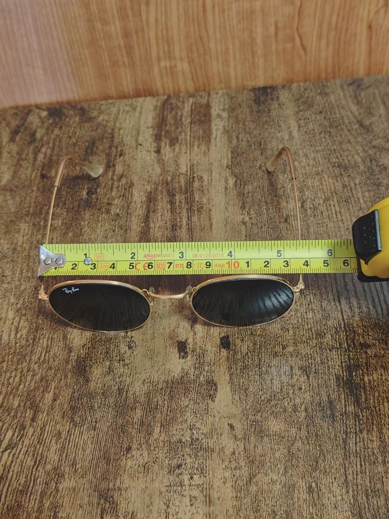May include: Gold-framed sunglasses with dark lenses, measuring approximately 5.5 inches in width. The Ray-Ban logo is visible on the left lens. The glasses are set against a wooden background, with a measuring tape indicating the size.