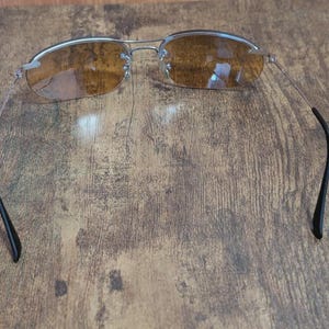 May include: Rimless sunglasses with a silver frame and brown-tinted lenses. The temples are thin and silver, curving into black earpieces. The glasses are set against a wooden background.