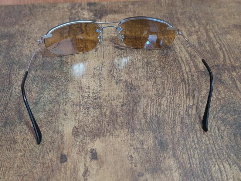 May include: Rimless sunglasses with a silver frame and brown-tinted lenses. The temples are thin and silver, curving into black earpieces. The glasses are set against a wooden background.