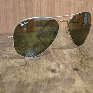 May include: Aviator-style sunglasses with a gold-colored metal frame and dark lenses. The Ray-Ban logo is visible on the upper left lens. The sunglasses are resting on a wooden surface.
