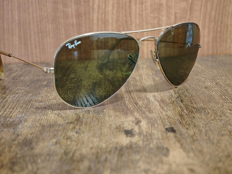 May include: Aviator-style sunglasses with a gold-colored metal frame and dark lenses. The Ray-Ban logo is visible on the upper left lens. The sunglasses are resting on a wooden surface.