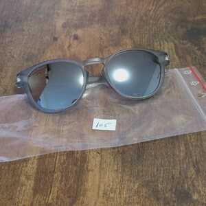 May include: Gray round sunglasses with dark lenses rest on a clear plastic bag. The sunglasses have a matte finish and a keyhole bridge. A small white tag with the number "105" is visible.